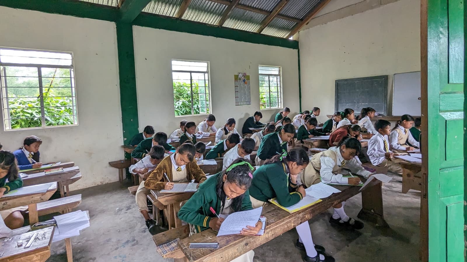Students in green and white uniforms writing at wooden desks in a tin-roofed classroom at Seng Khasi School, Ri-Bhoi District, Meghalaya