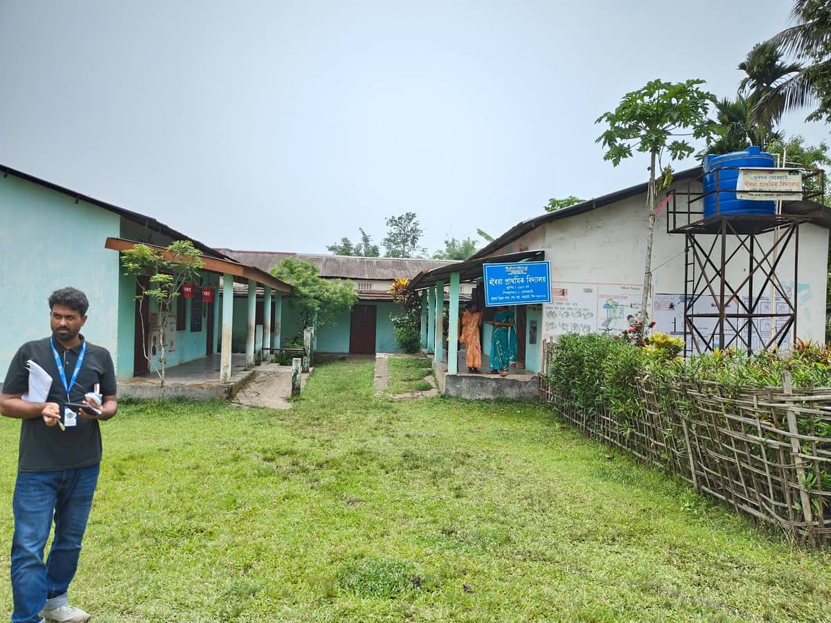 A field team member holding notes in the grassy courtyard of Rowriah LP School, Jorhat, Assam
