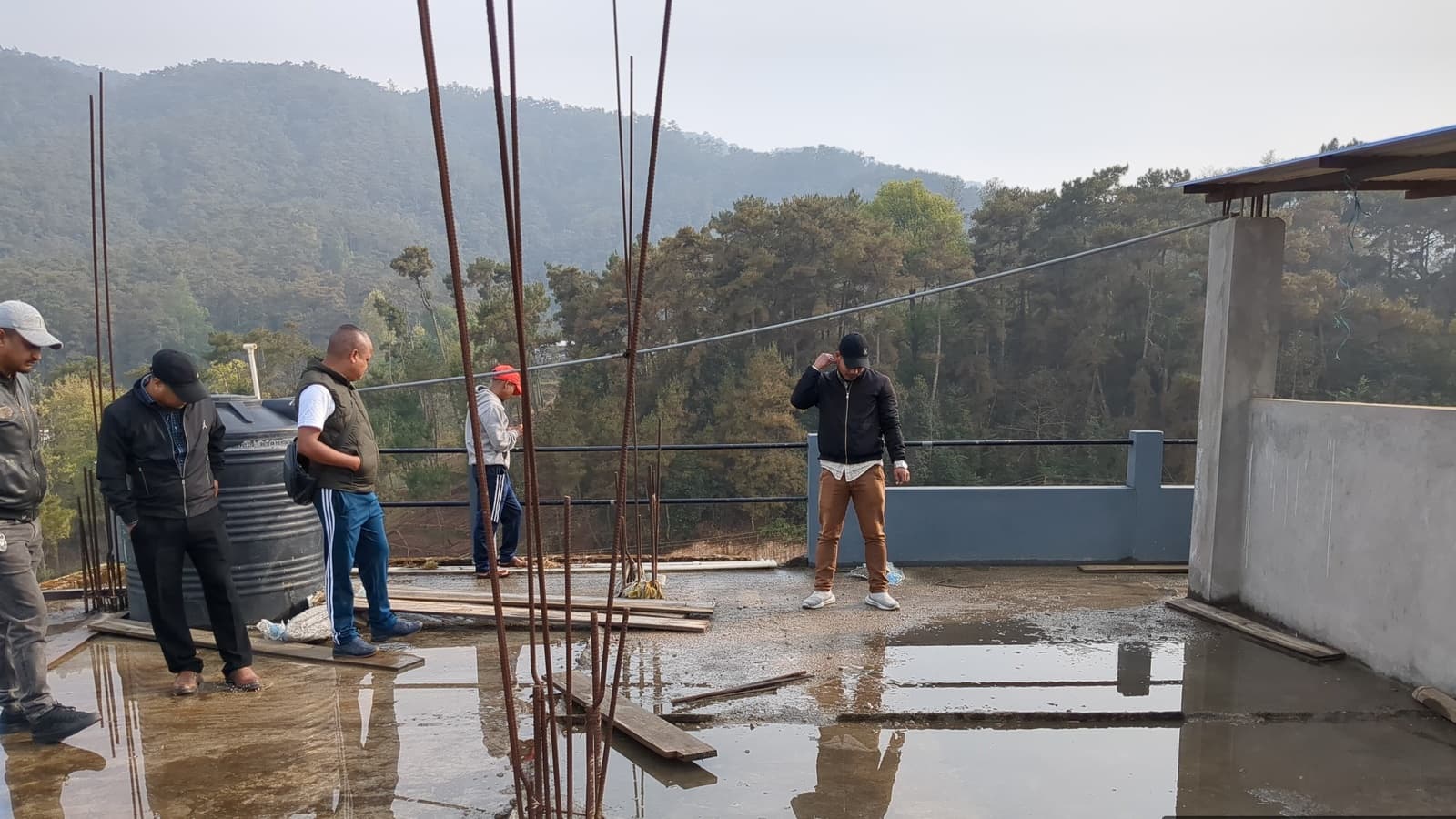 The field team walking an unfinished rooftop with steel rods and forested hills behind, at SOSO Tham Senior Secondary School, Lawsohtun, Meghalaya