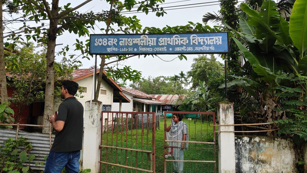 A field team member at the gate of Nowchaliyan Gaon LP School, Jorhat, Assam, under the school's blue signboard