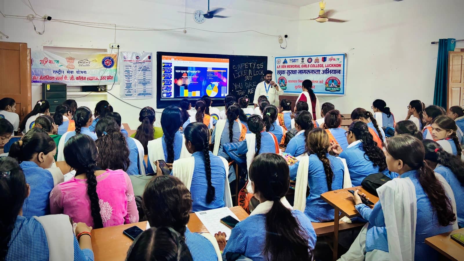 A facilitator holding a microphone beside a screen, facing a full classroom of students at A.P. Sen Memorial Girls College, Lucknow